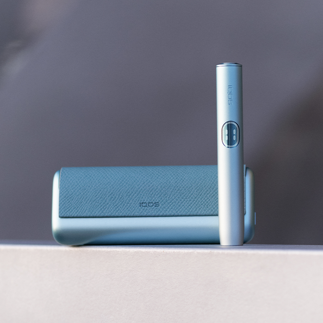 IQOS ILUMA i PRIME Garnet krāsā atrodas uz koka galda izplūdušā fonā.