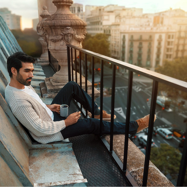 Мужчина сидит на террасе с чашкой кофе и видом на город.